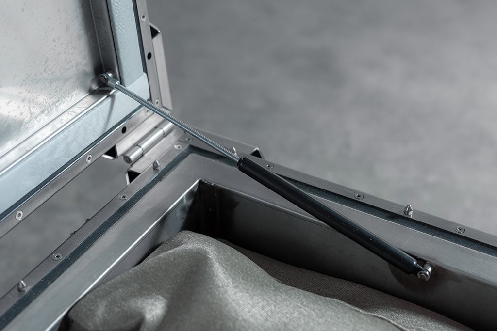 Una scatola metallica aperta con molla a gas nera e rivestimento in tessuto grigio.