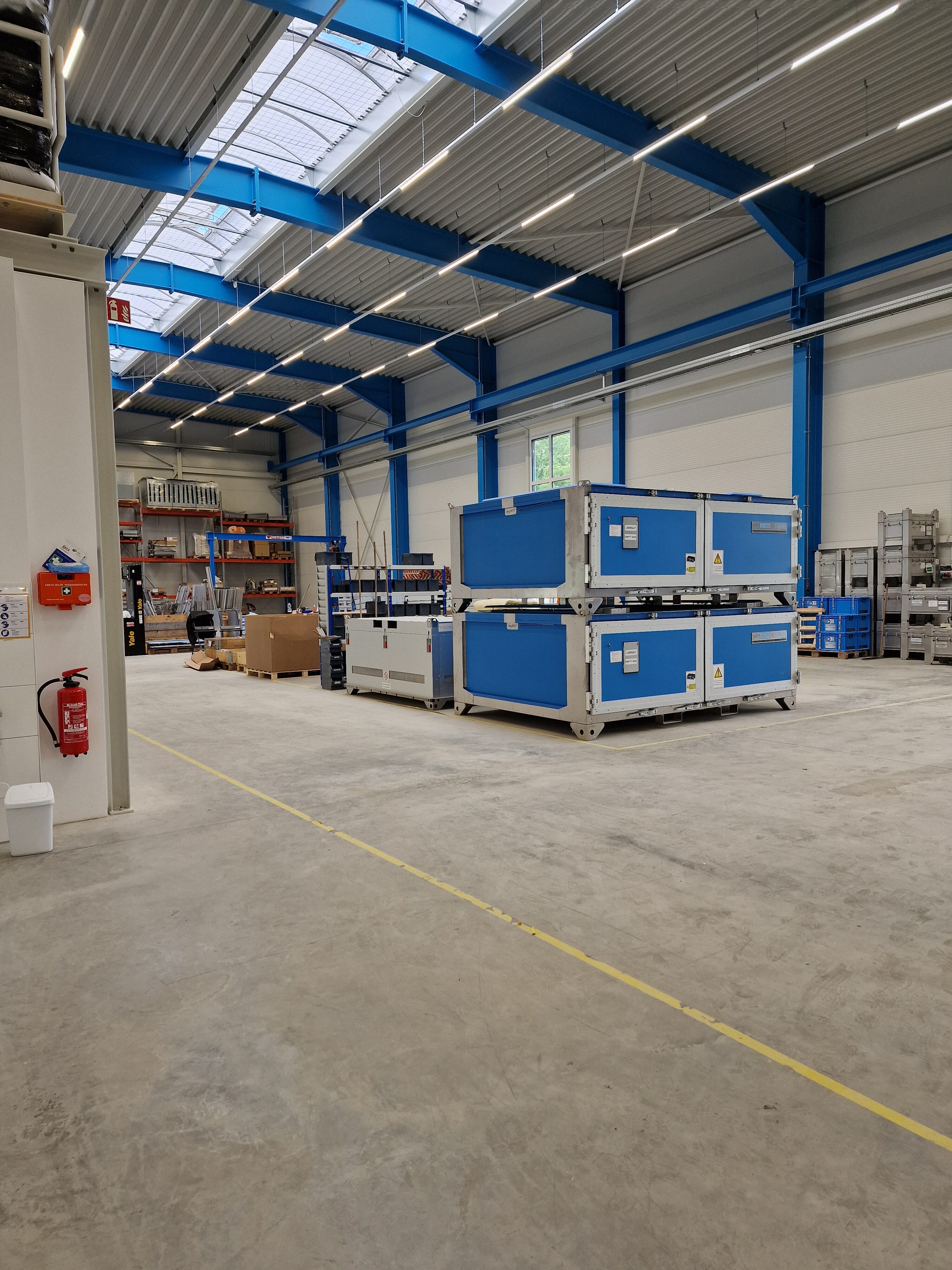 Grande magazzino con container blu, scaffali e scatole di cartone; spazio libero sul pavimento. Illuminazione intensa.