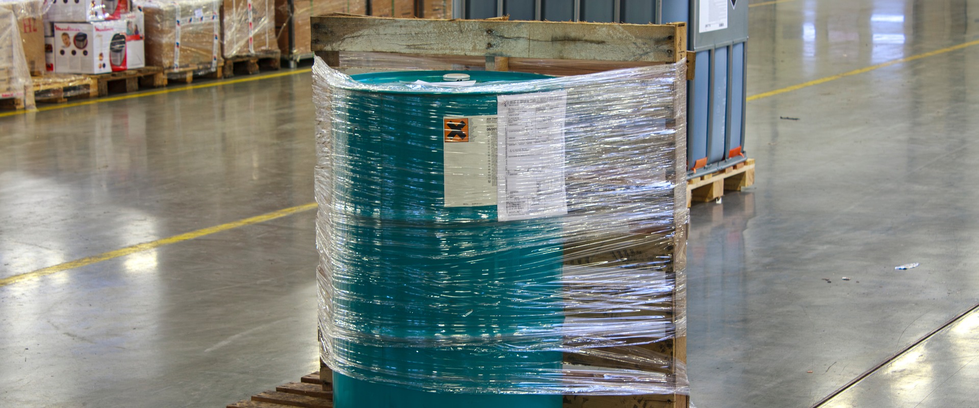 Botte verde avvolta nella plastica e fissata su un pallet di legno. La botte è circondata da merci imballate e da un altro grande container, il che fa pensare a un ambiente di stoccaggio ben organizzato.